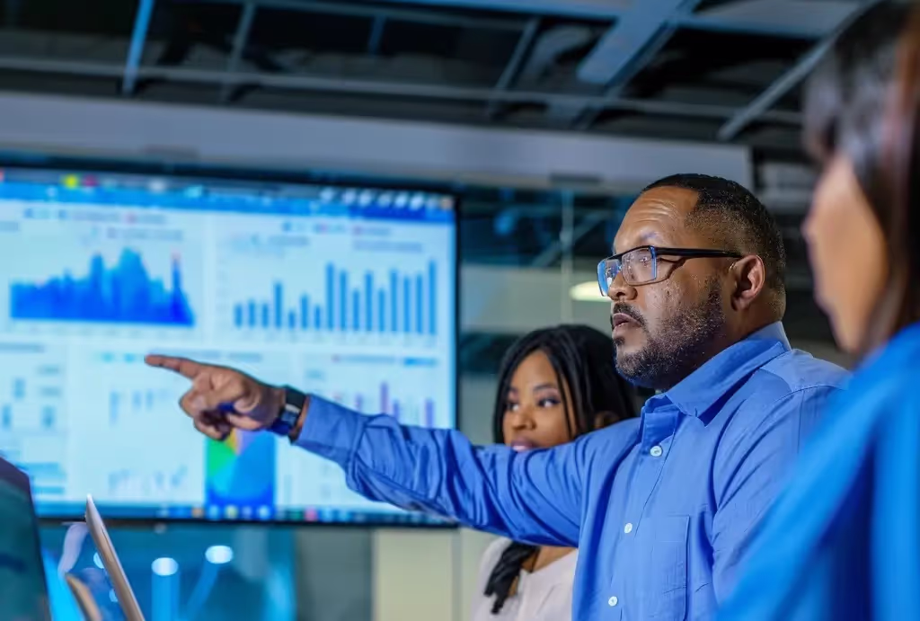 Man pointing at a large screen of bar charts during a team briefing